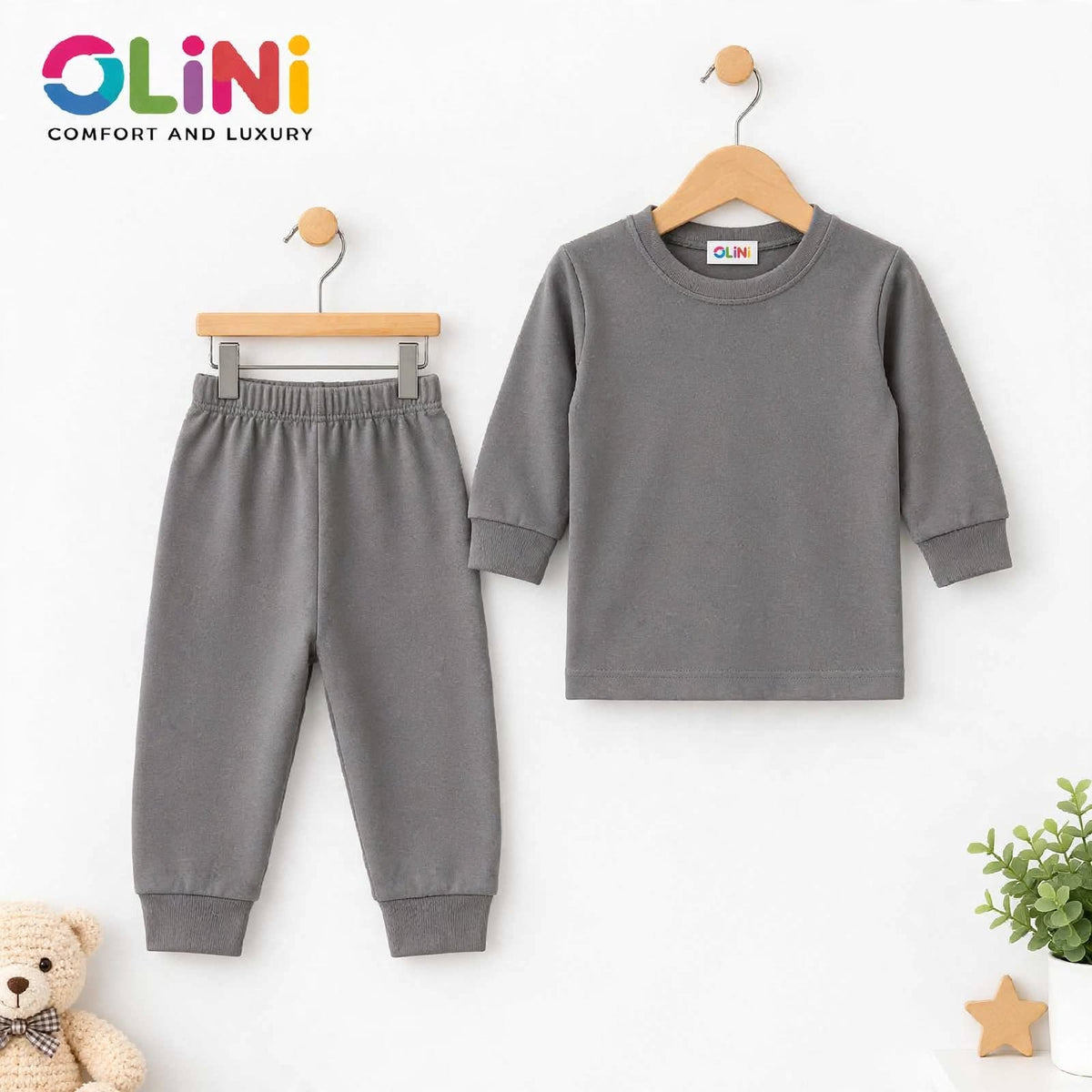 A two-piece children's loungewear set by OLiNi, featuring a long-sleeve gray top and matching jogger pants, displayed on wooden hangers against a clean white background with a teddy bear and small plant props.