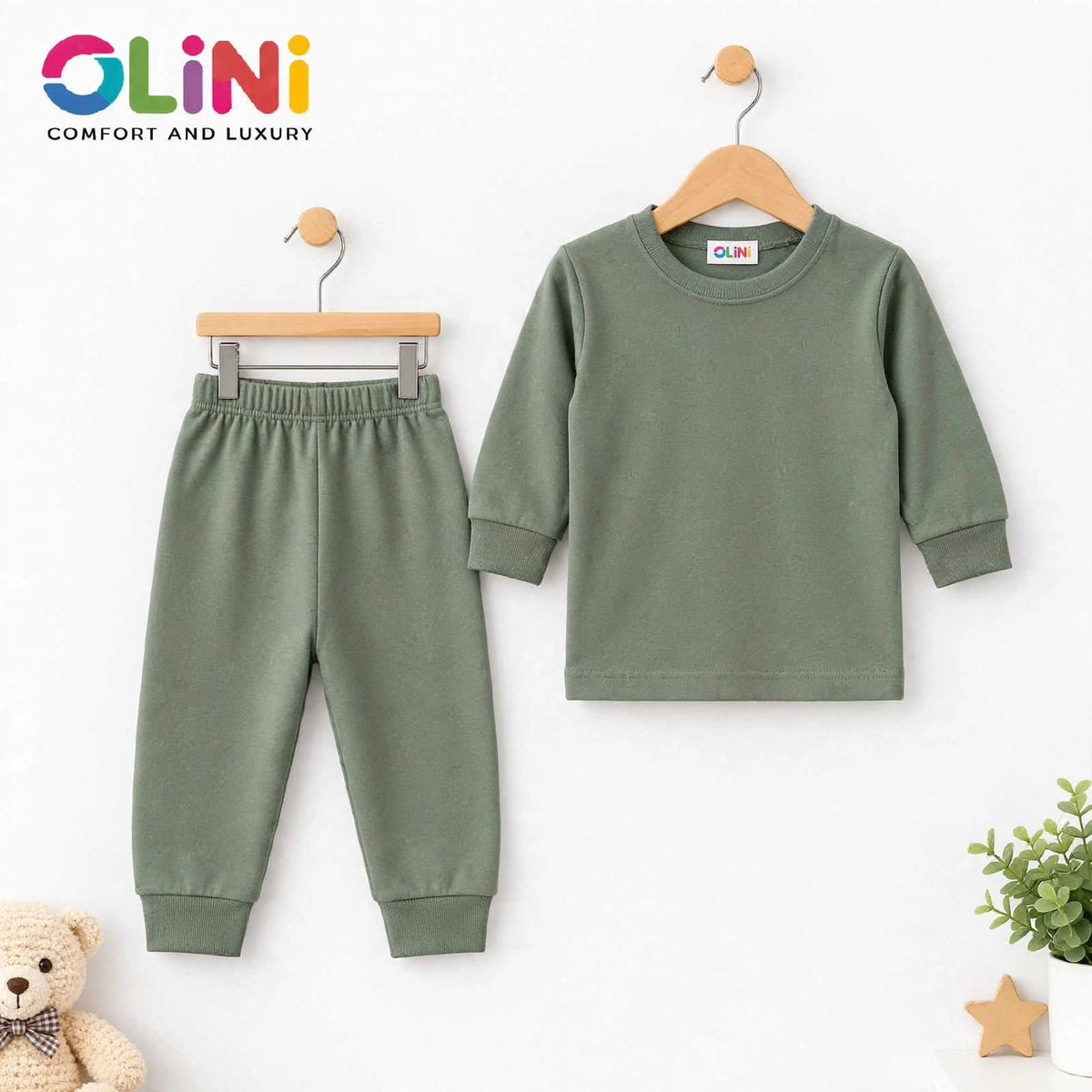 A two-piece children's loungewear set by OLiNi, featuring a long-sleeve gray top and matching jogger pants, displayed on wooden hangers against a clean white background with a teddy bear and small plant props.